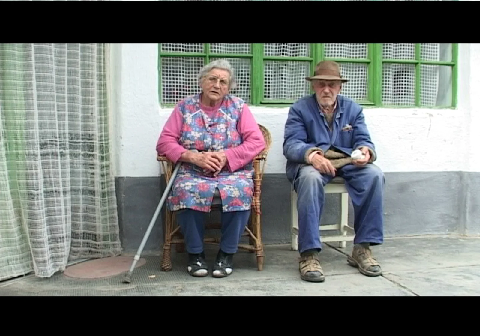 An older woman and man sit outside a house in The Angelmakers.